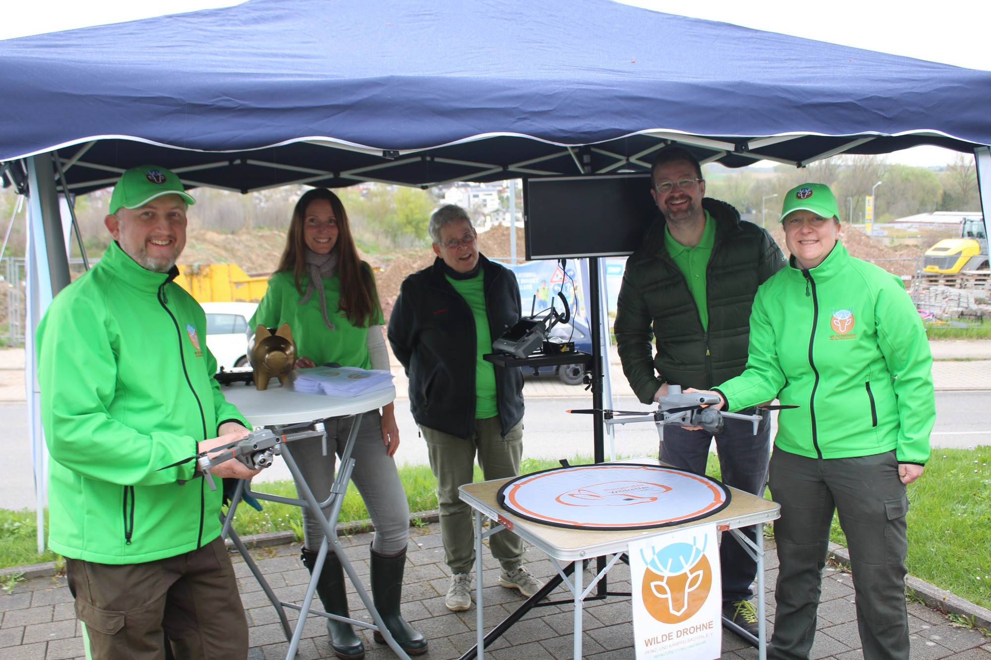 Gruppe von fünf Personen in grüner Kleidung unter einem Zelt, verschiedene Aktivitäten.