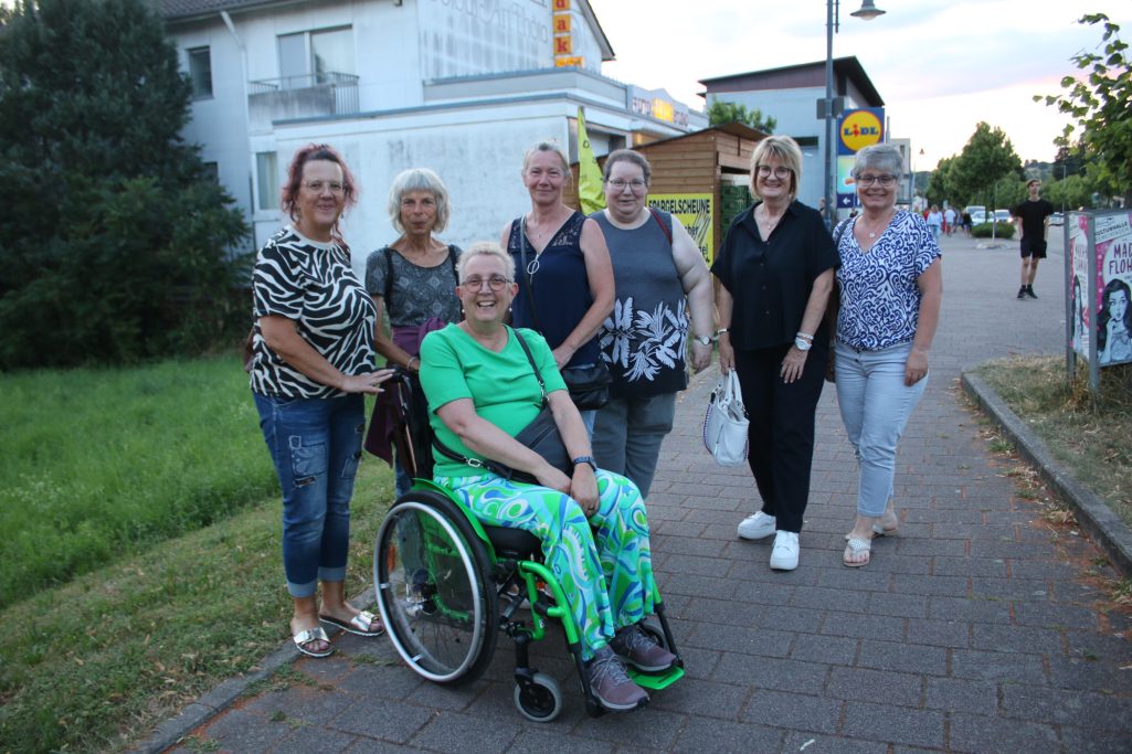 Gruppe von sieben Frauen, darunter eine im Rollstuhl, stehen auf einem Gehweg.