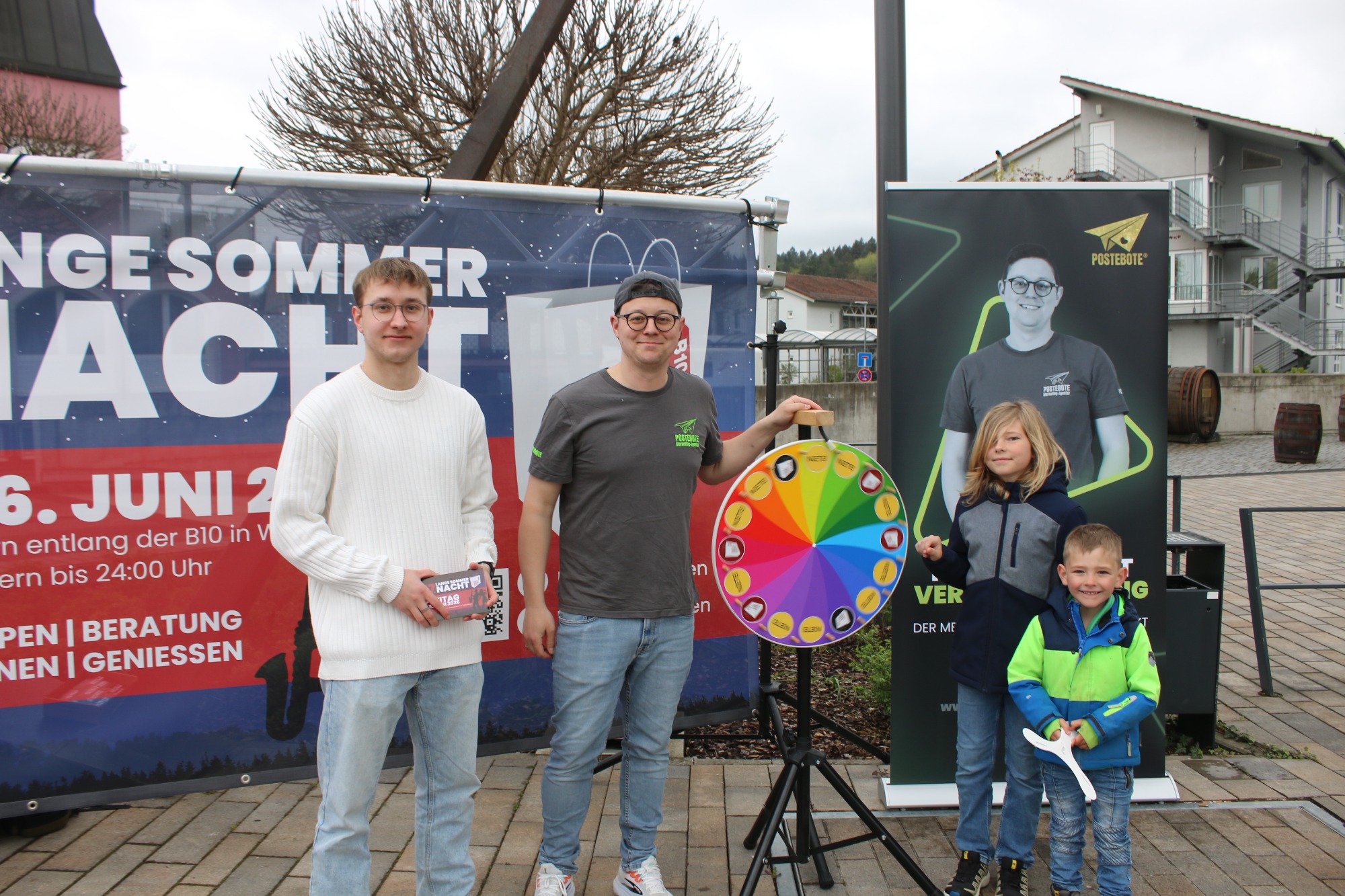 Zwei Männer und zwei Kinder stehen vor einem Glücksrad und einer Werbefläche.