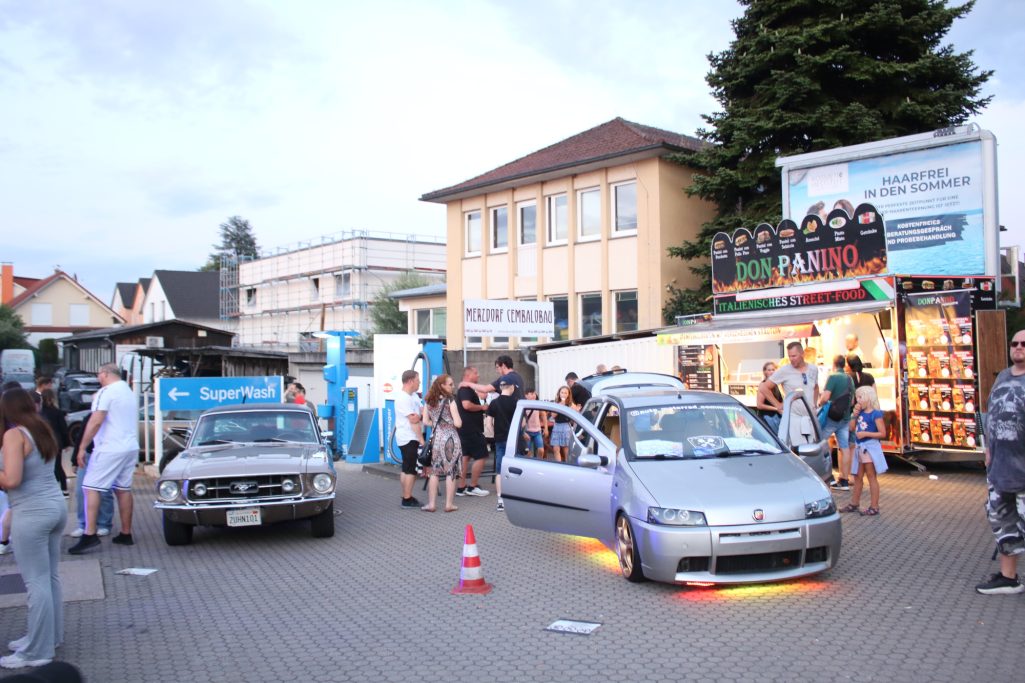 Menschenmenge auf einem Parkplatz mit Autos und Verkaufsständen bei Abendlicht.