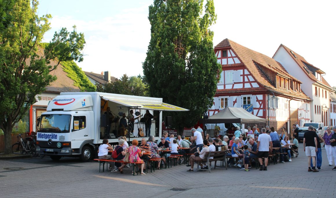 Menschen sitzen draußen an Tischen, während ein Foodtruck und eine Bühne sichtbar sind.