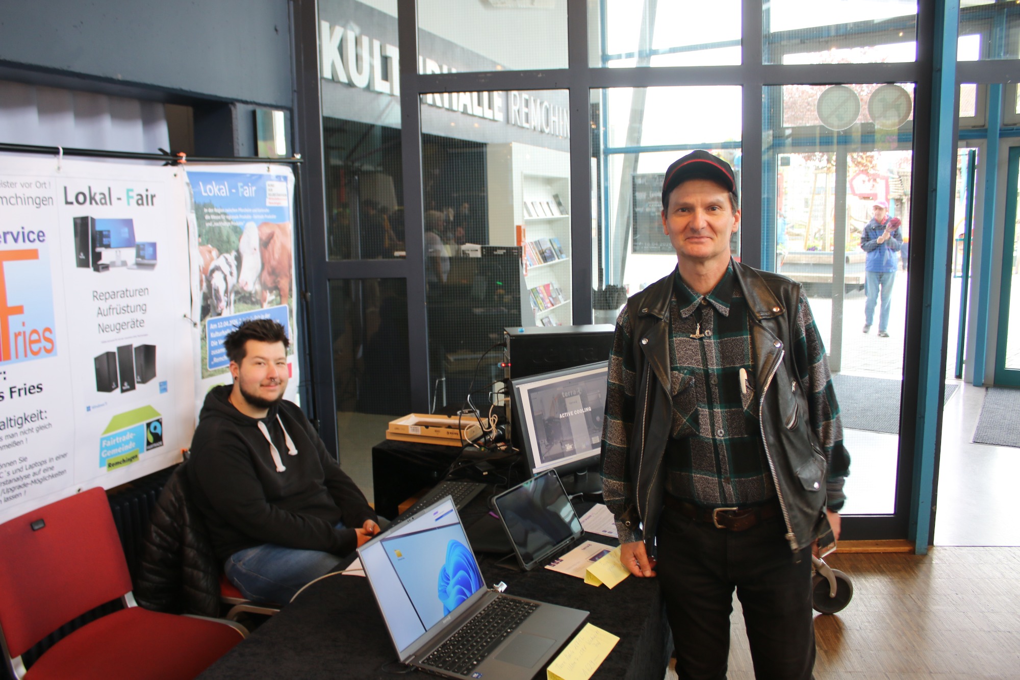 Zwei Männer stehen an einem Tisch mit Laptops und Informationsmaterialien.