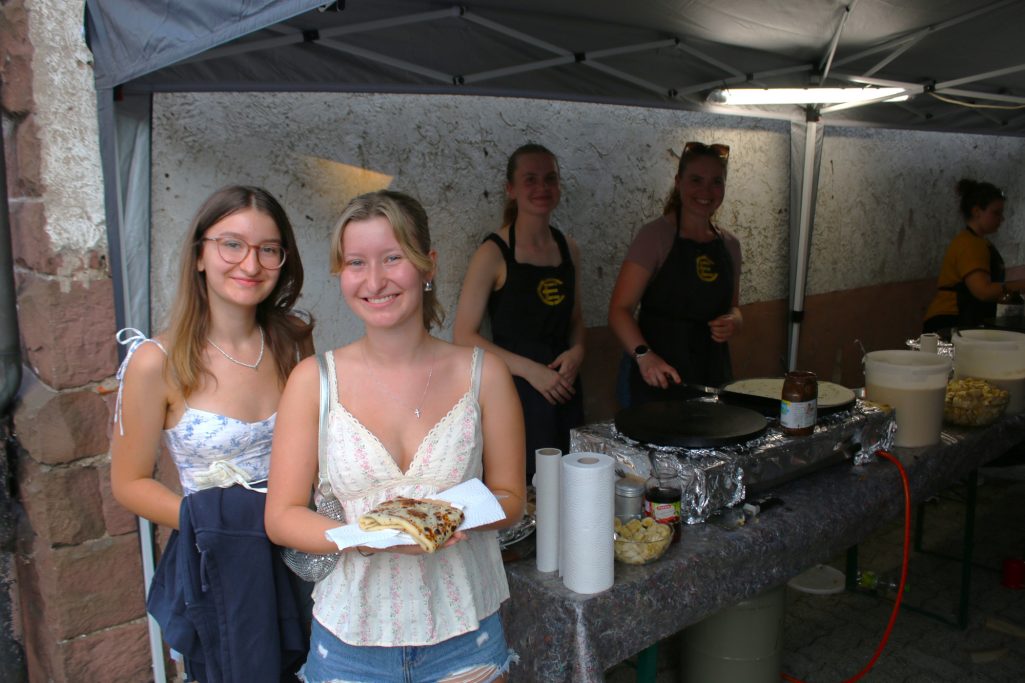 Zwei junge Frauen lächeln, während im Hintergrund Verkäufer am Grillen sind.