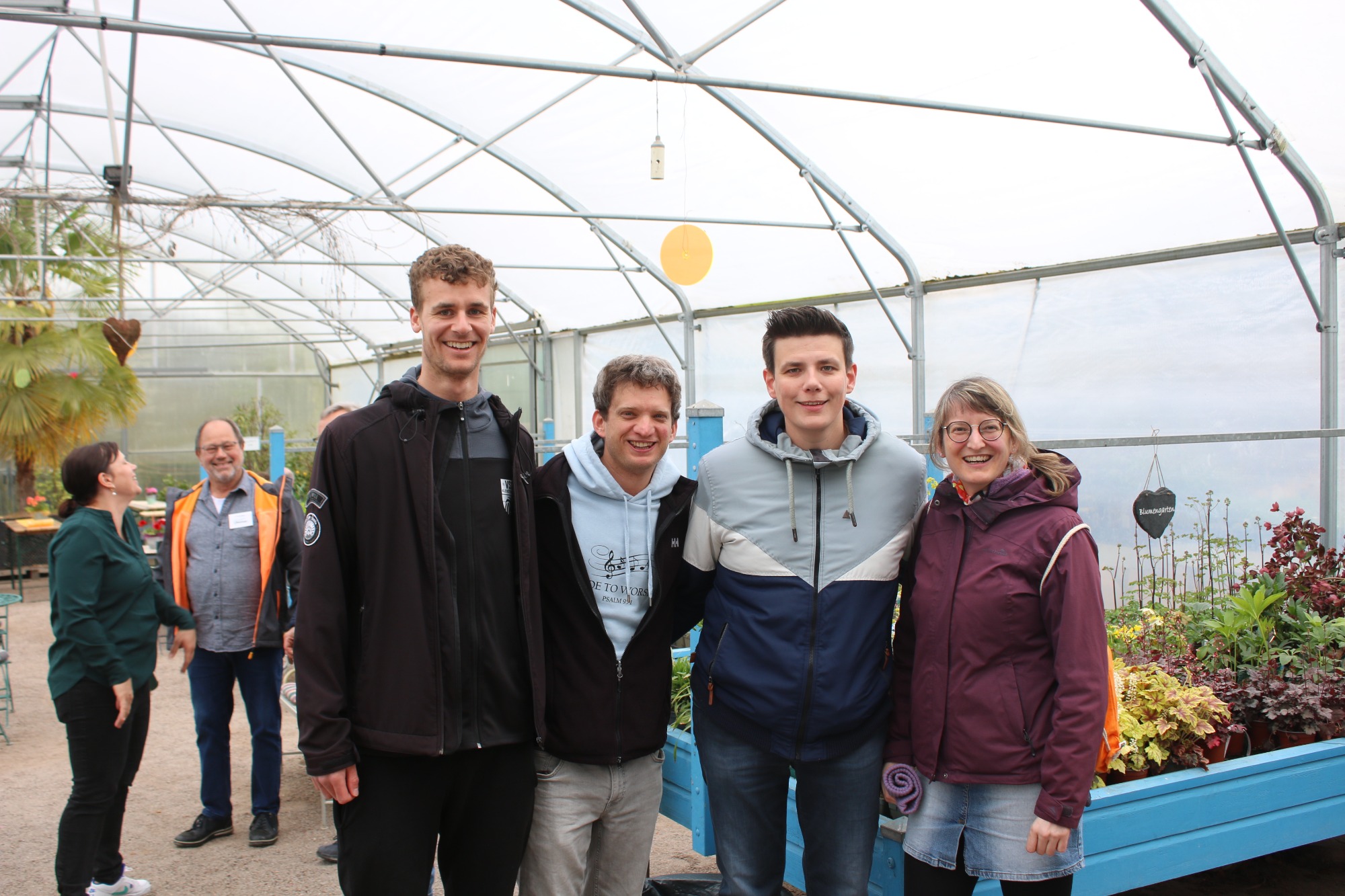 Vier Personen in einem Gewächshaus, umgeben von Pflanzen und bunten Blumen.