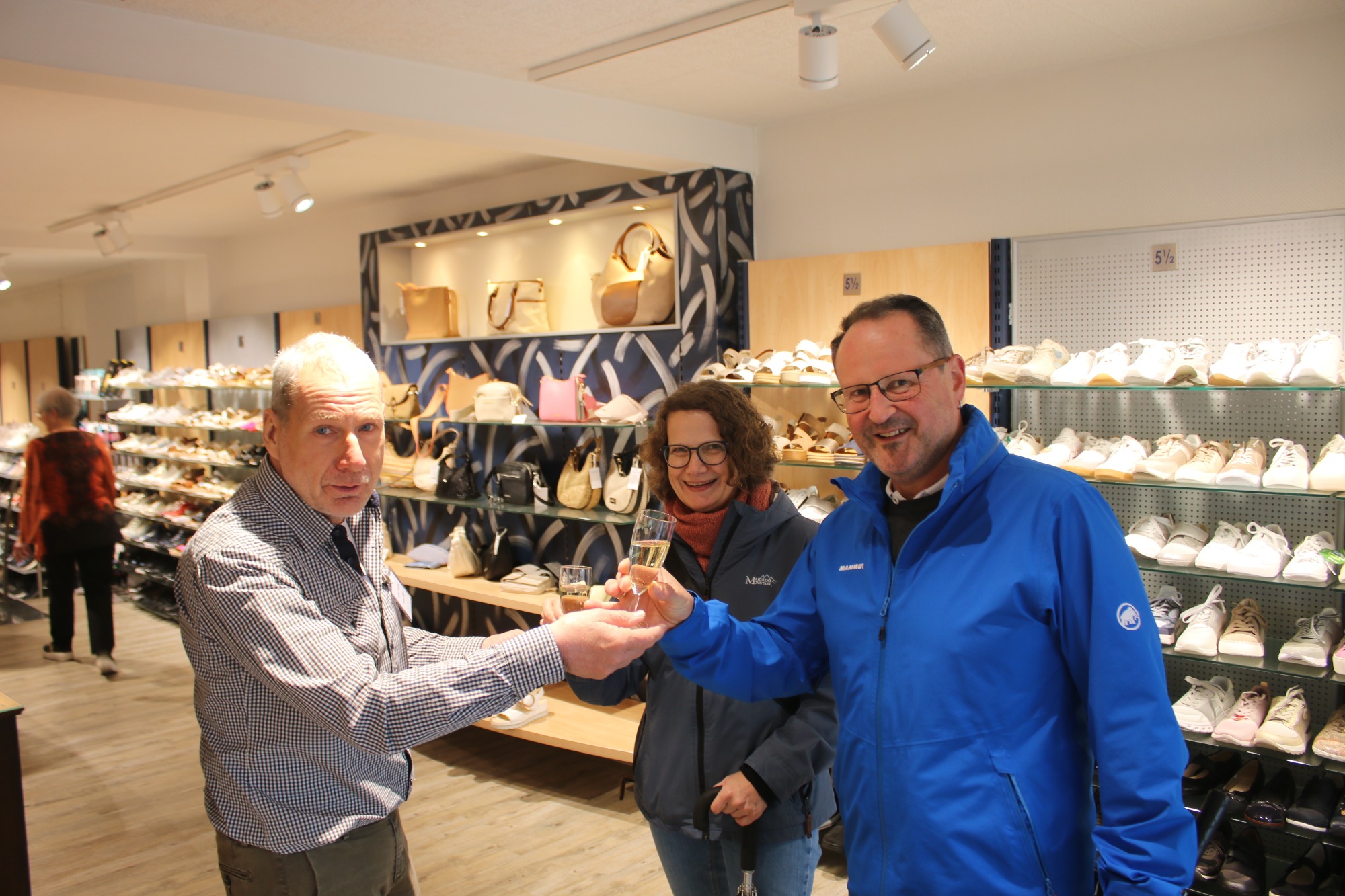 Drei Personen in einem Schuhgeschäft, lächelnd und einen Gegenstand händeschüttelnd.