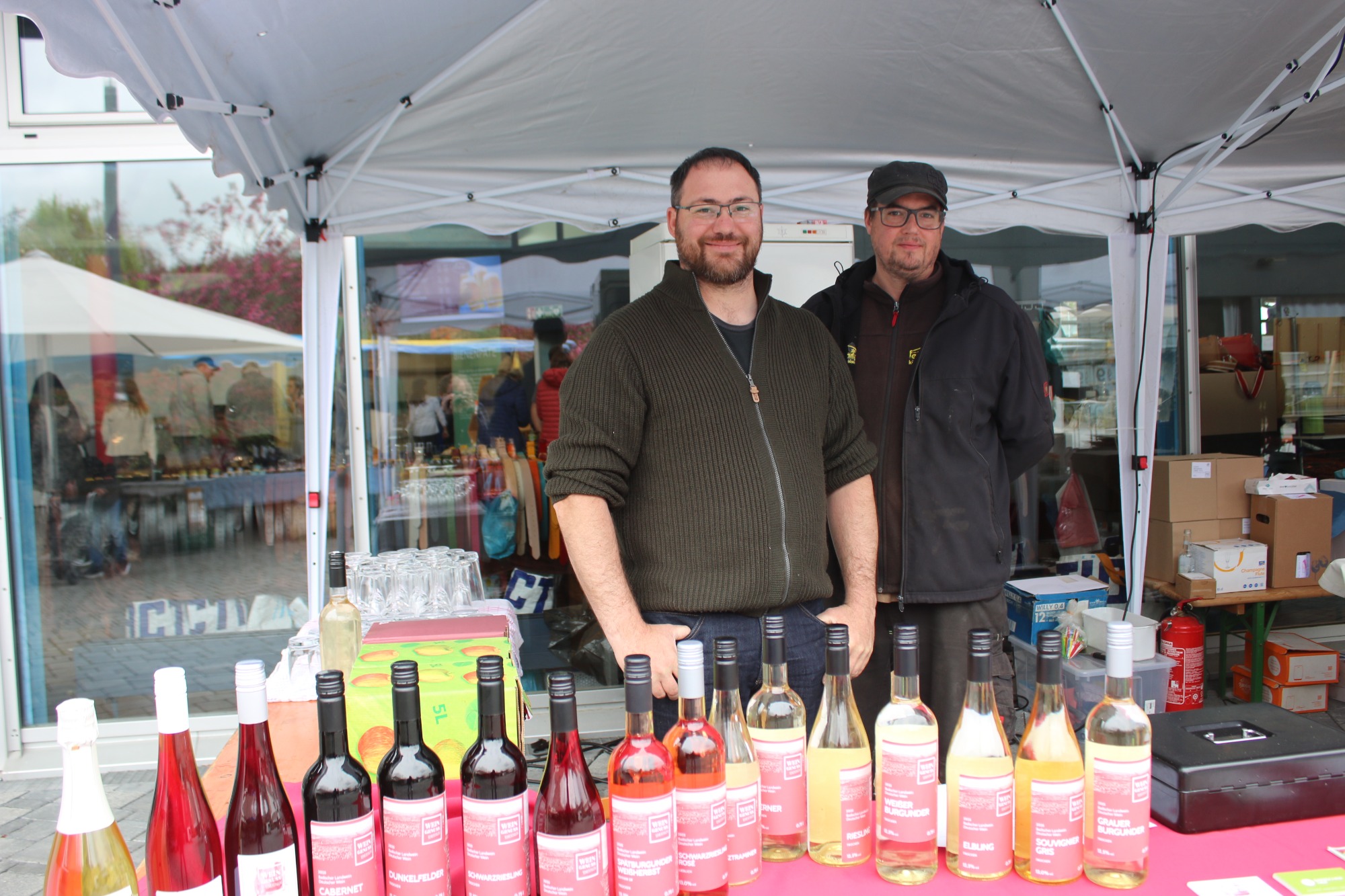 Zwei Männer stehen hinter einem Tisch mit verschiedenen Weinflaschen auf einem Markt.