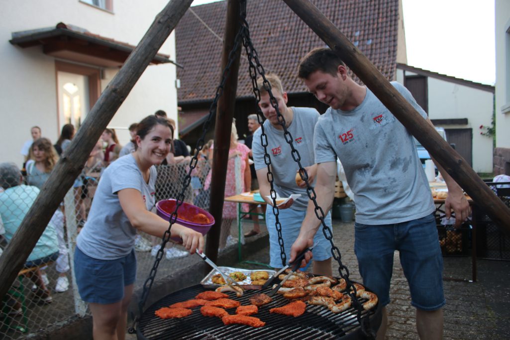 Drei Personen grillen Würstchen und Gemüse auf einem großen Grill bei einer Feier.