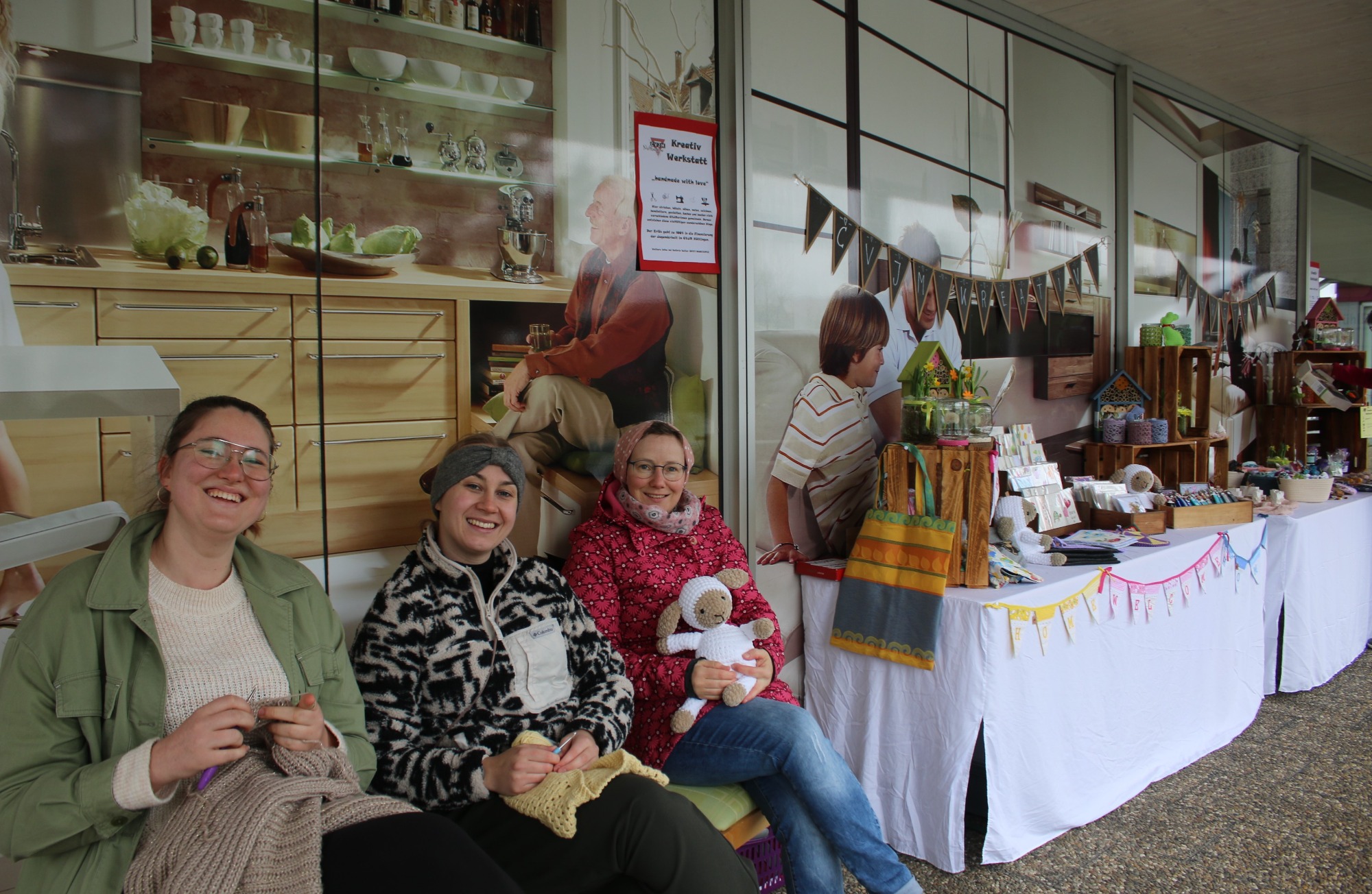 Drei Personen sitzen vor einem Marktstand mit handgefertigten Produkten.