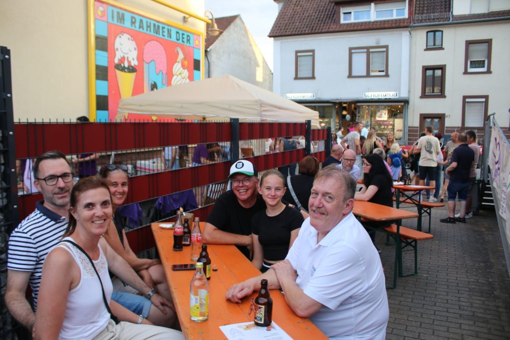 Gruppe von Menschen sitzt an Tischen in einer belebten Straße, Getränke vor ihnen.