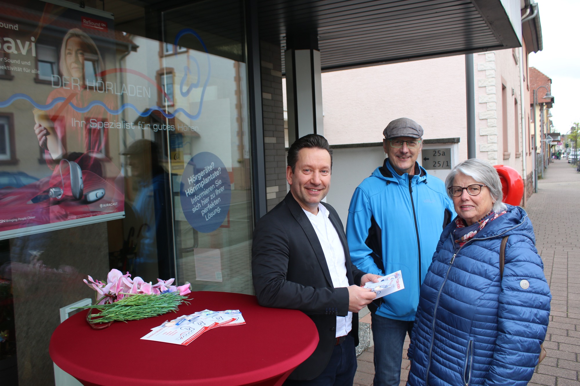 Personen stehen vor einem Geschäft, verteilen Informationsmaterial und lächeln freundlich.