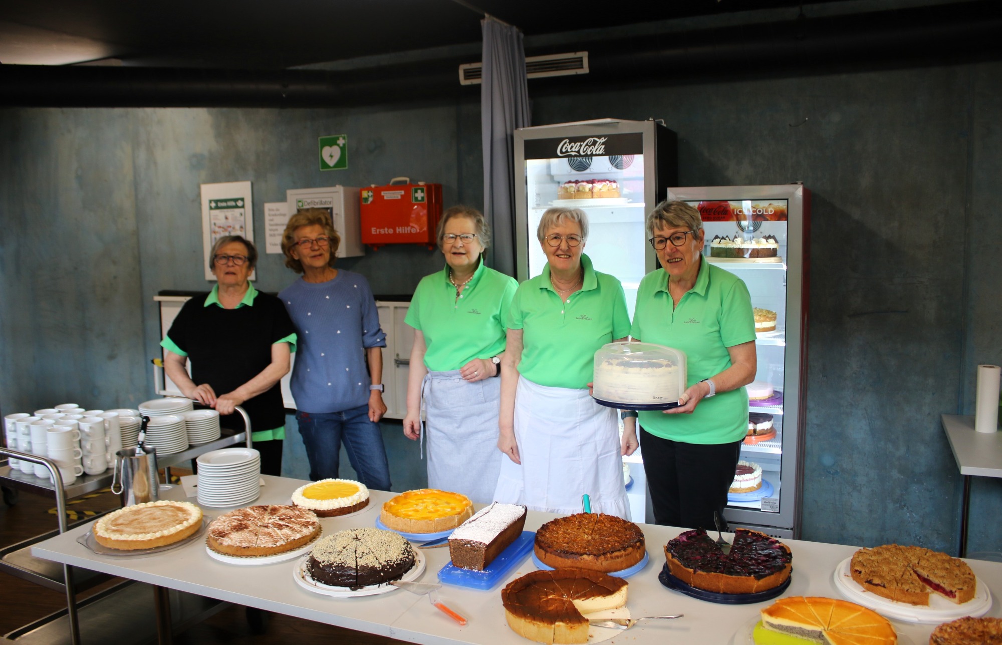 Fünf Frauen in grünen Shirts stehen hinter einem Tisch voller Kuchen und Torten.