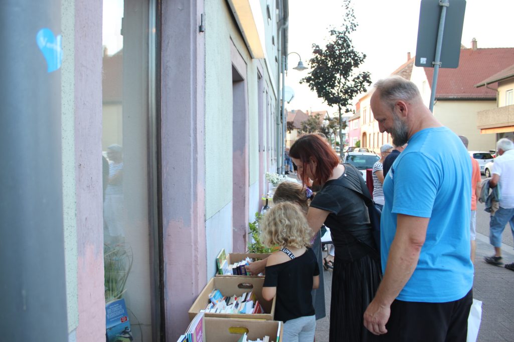 Eine Familie mit Kindern schaut sich Produkte in Kisten an, die vor einem Gebäude stehen.