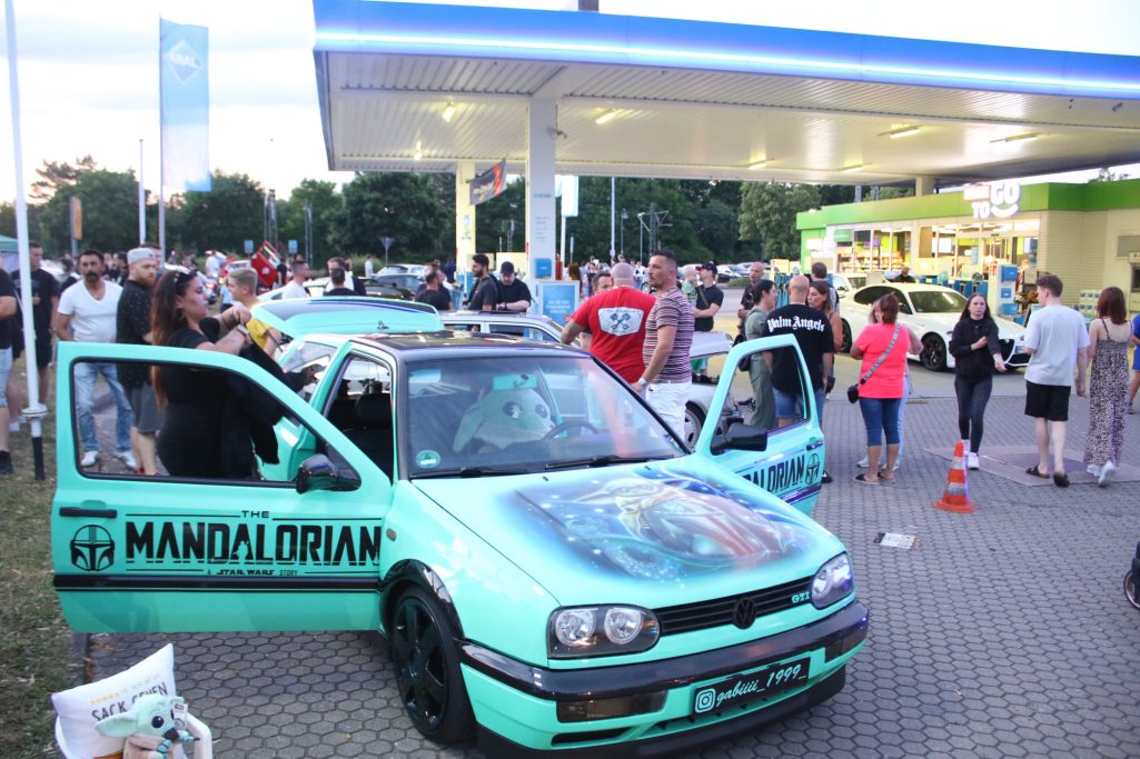 Personen versammeln sich um ein modifiziertes Auto mit auffälliger Lackierung an einer Tankstelle.