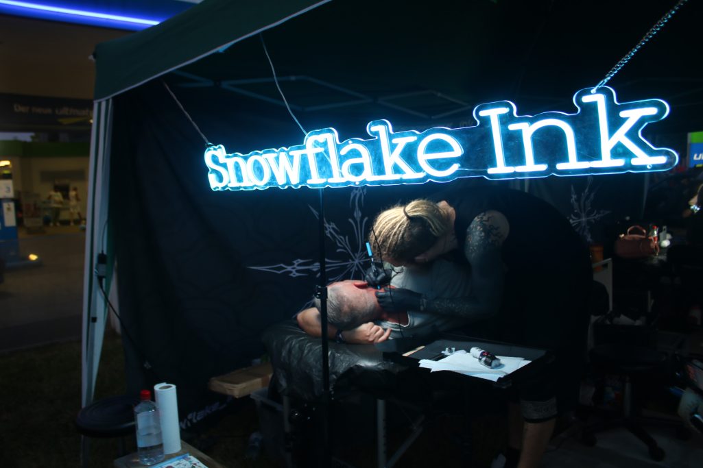 Stand von "Snowflake Ink" mit neonblauer Beleuchtung und Tätowierer bei der Arbeit.