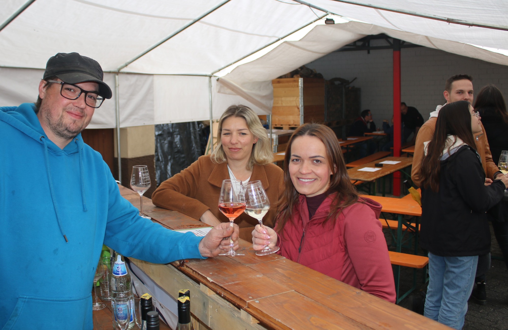 Drei Personen stoßen mit Gläsern in einem Zelt an, im Hintergrund weitere Gäste.