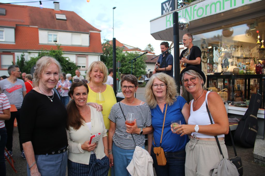 Gruppe von sechs Frauen lächelt vor einem Geschäft auf einer belebten Straße.
