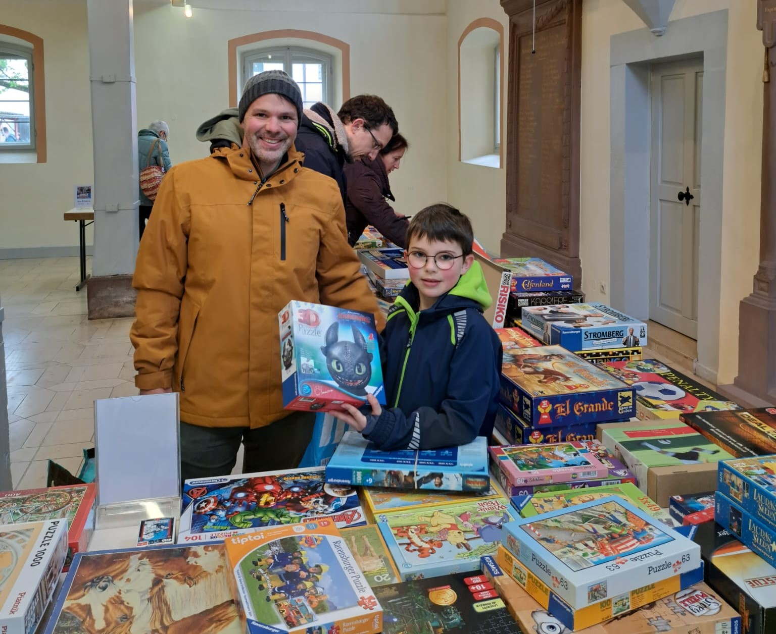 Zwei Personen lächeln vor einem Tisch voller Brettspiele und Puzzle.