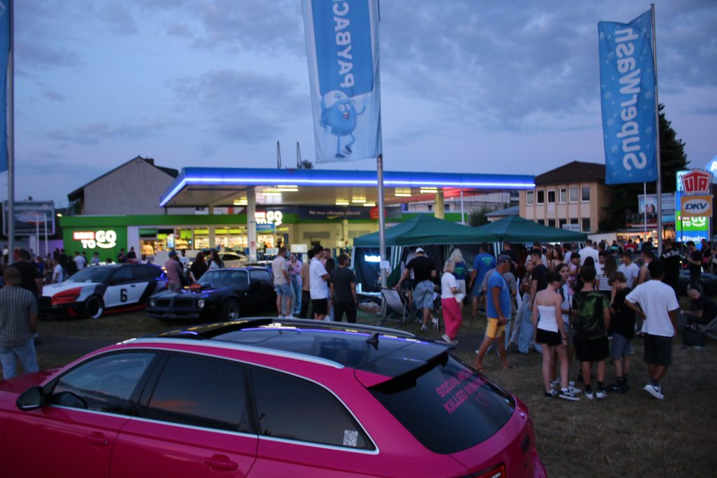Bunte Menschenmenge bei einem nächtlichen Autotreffen vor einem Geschäft.